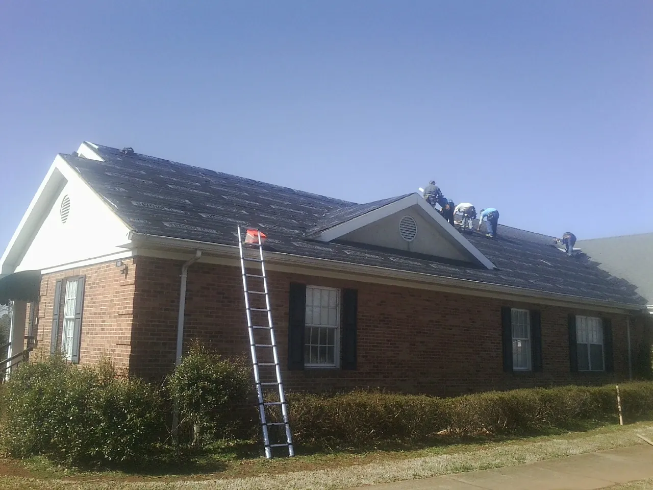 Crew tearing shingles off a brick ranch roof with a ladder set — Emergency Roof Repair work by our crew in Carriere