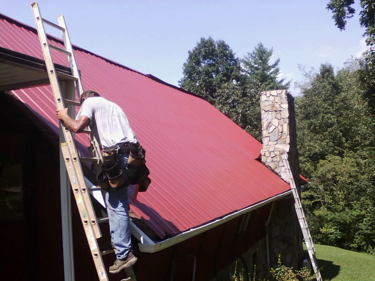 Roofer on a ladder beside a red metal roof and stone chimney — Soffit & Fascia Repair by Roofing Brothers in Carriere